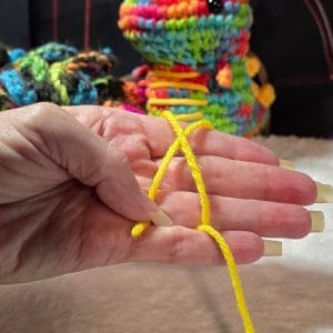 Hand demonstrating the starting loop of a magic circle with yellow yarn, shown against a soft background. Long, manicured nails and colorful plushies in the backdrop add a playful, comfort-core aesthetic—ideal for beginners learning how to make a magic circle in crochet.