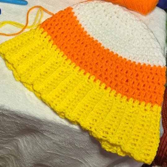 A close-up of a crochet beanie in candy corn colors with visible ribbed yellow brim, displayed on a white surface with nearby crochet tools and yarn.