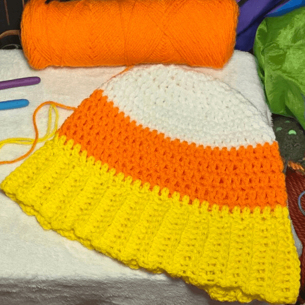 In-progress Candy Corn Beanie crochet project with white, orange, and yellow stripes, displayed beside orange yarn and crochet hooks.