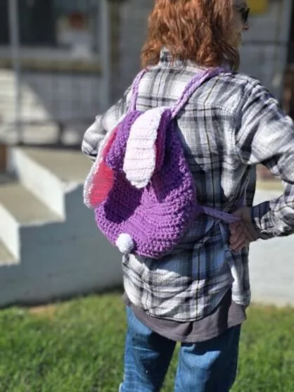 Person standing on a grassy lawn wearing a handmade purple crochet bunny backpack with white and pink ears and a fluffy pompom tail; whimsical, cozy, and child-friendly design.