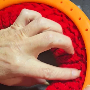 Close‑up of a loom knitter using a pink‑handled hook to lift red yarn loops over the pegs on an orange knitting loom.