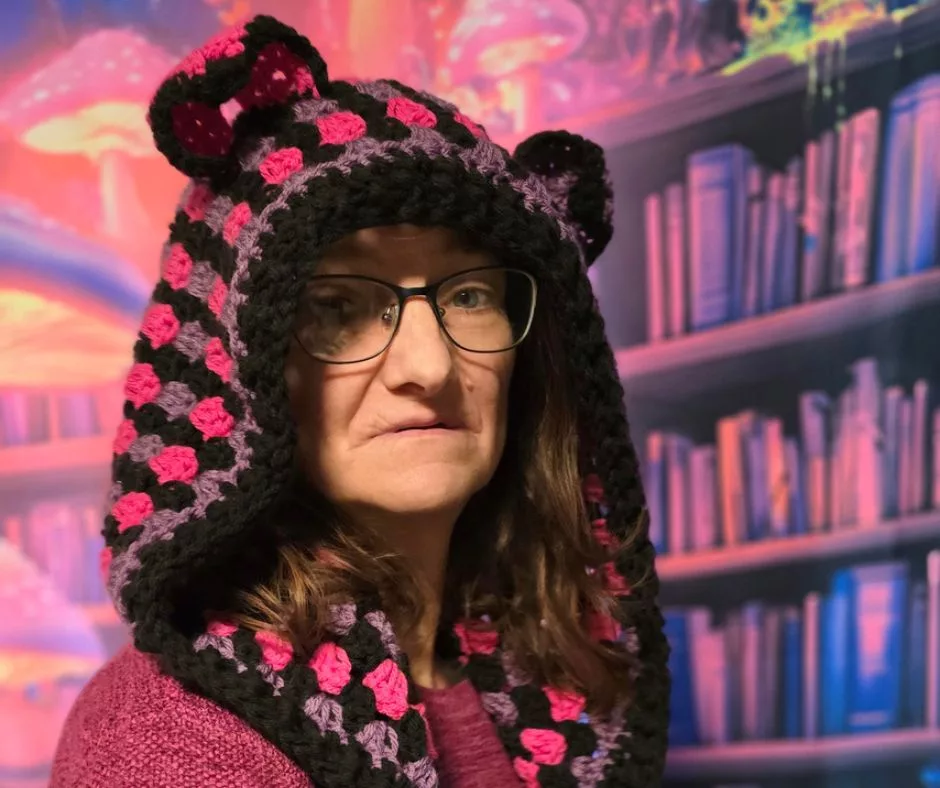 A person wearing glasses and a colorful crochet hood with cat ears in black, pink, and purple yarn. The background is a vibrant fantasy scene with glowing mushrooms and bookshelves.