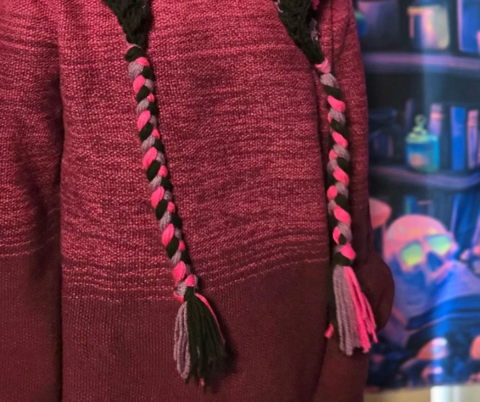 Close‑up of braided tassels on a handmade crochet hood in black, pink, and gray yarn. The wearer has on a maroon sweater, and the background features colorful fantasy shelves with glowing bottles.