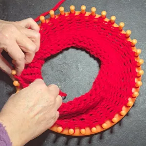 Hands working on a round knitting loom with red yarn, showing the knitted fabric forming as loops are lifted over the pegs.