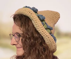 A side profile of a woman with long, curly hair wearing a handmade, tan crocheted hedge witch hat. The hat features a ruffled brim and is adorned with small, dark blue and green crocheted flowers around the base of the crown.
