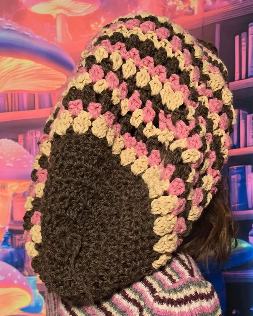 A person wearing a large Neapolitan‑colored crochet beanie in brown, pink, and Buff pulled playfully over their head, with a fantasy backdrop of oversized colorful mushrooms and bookshelves.