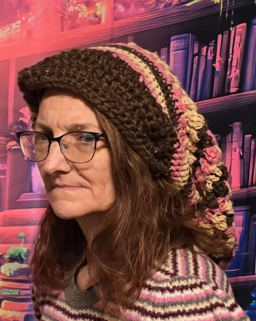 A person wearing glasses and an easy slouchy beanie crochet pattern in brown, pink, and Buff, standing in front of vibrant bookshelves lit with pink and purple light.