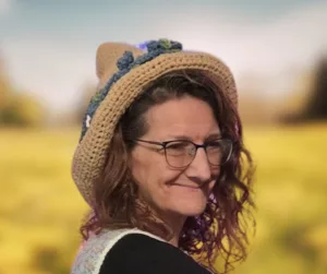 A close-up, slightly tilted view of a tan crocheted hedge witch hat with a whimsical pointed top. The design includes a textured crochet pattern and a row of blue and green floral embellishments along the brim's edge.