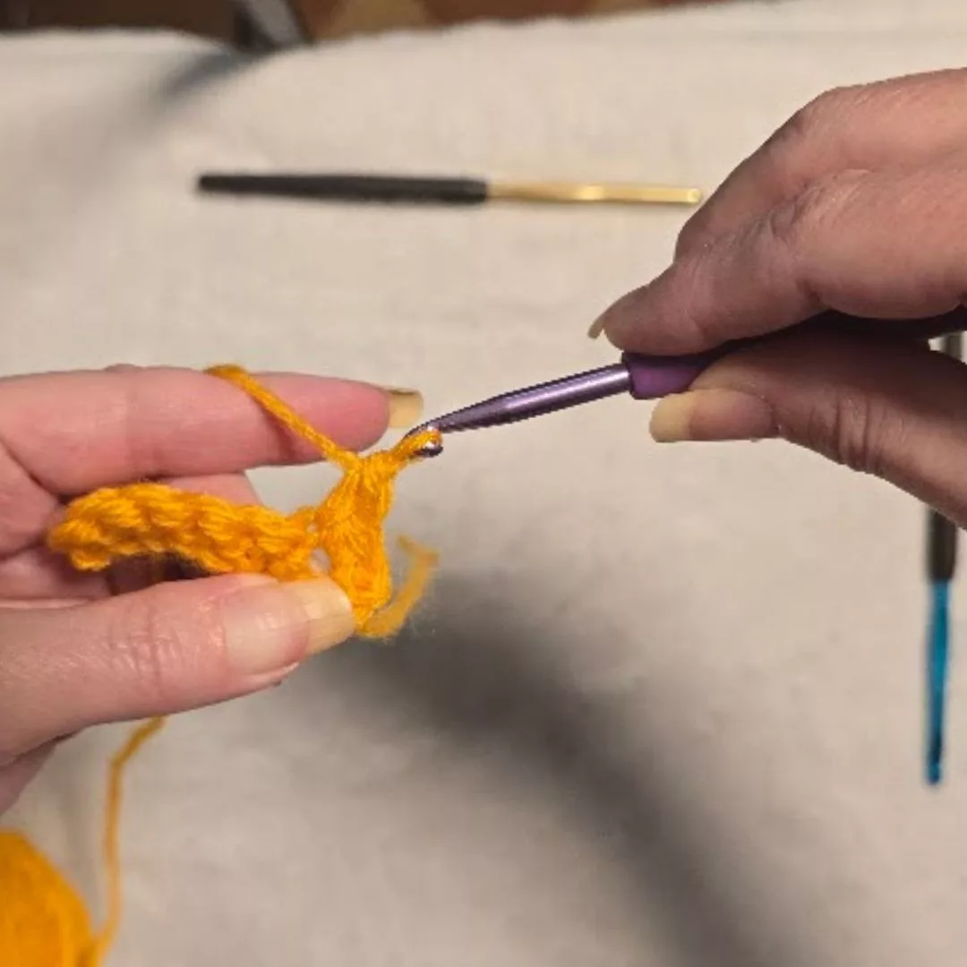 Close-up of the final "pull through" step for a single crochet: The blue-handled hook catches the yellow yarn and is pulled through both remaining loops on the hook. This action clears the hook of all but one loop, completing the stitch and securing the fabric.