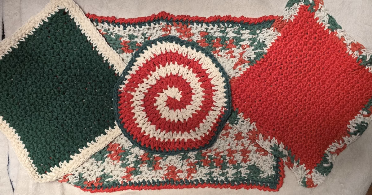 Four-piece Christmas crochet kitchen set laid out with red, green, and ivory cotton dishcloths, a dishtowel, and a peppermint spiral trivet.